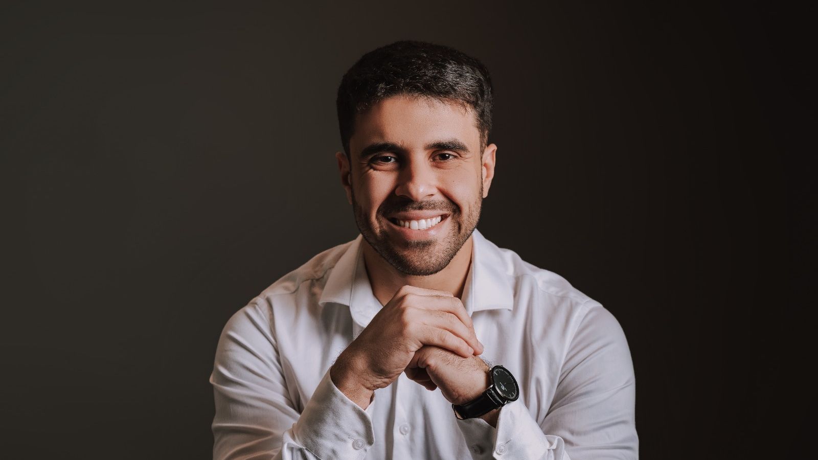Leonardo Ferreira Santos, psicólogo, de camisa branca, mãos entrelaçadas, sorrindo